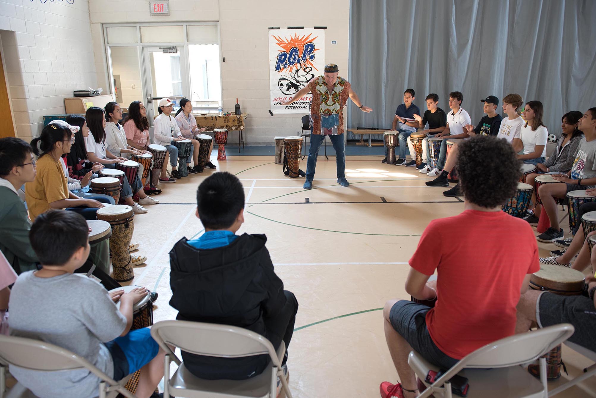 Students sitting in a circle with drumming teacher standing in the middle doing a lesson Open Gallery